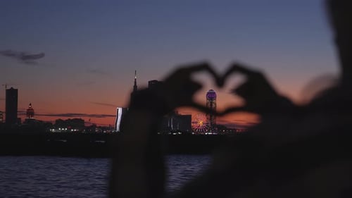 Mujer haciendo corazón con las manos con vista al brillante paisaje urbano al atardecer