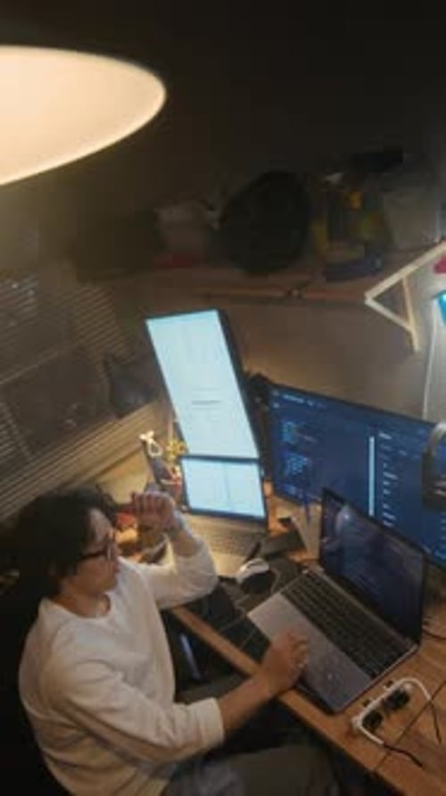 Man Coding on Laptop with Multiple Screens in Home Office at Night