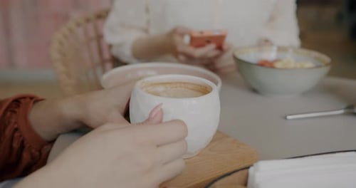 Closeup of Female Hands Holding Coffee Cup and Smartphone While Women Enjoying Drink in Cafe in
