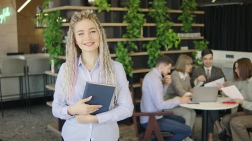 Young Woman with Tablet Smiling Confidently in a Modern Office Setting While a Group of Colleagues