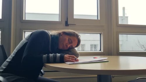 A woman is asleep at her office table, resting her head on her left arm, exhausted from the monotony