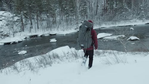 Traveler with Backpack Going Down Snowy Slope towards the River