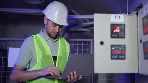 Electrical Engineer Works on Laptop Next to Control Panel in Industrial Setting