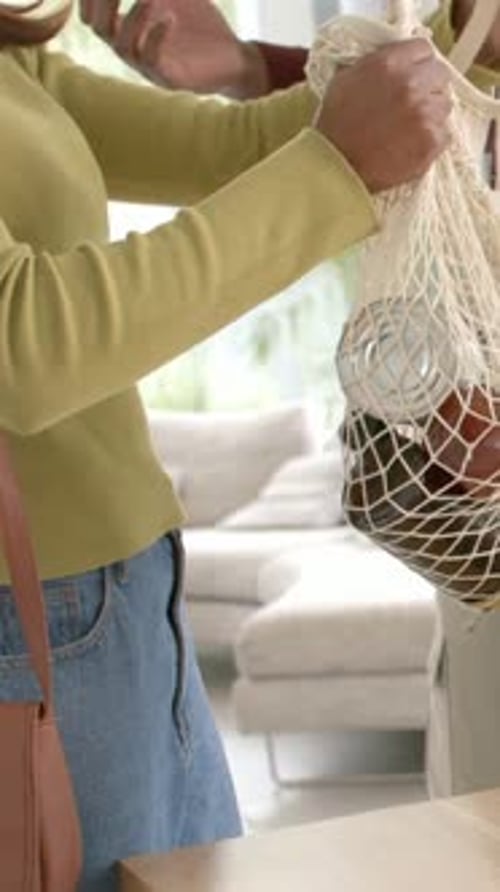 Vídeo vertical: Al entrar a casa, una mujer vestida con la parte superior verde con una bolsa de red colocando la comida en la mesa, espacio para copiar