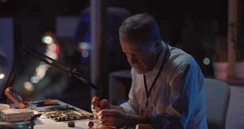Mature Development Engineer Sitting at Desk Soldering Microchip Circuit Board Looking to Camera