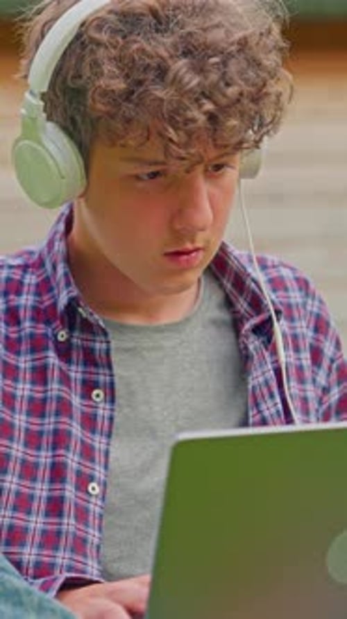 Vertical Video Curly Teenage Boy Listening to Music on Headphones and Works on a Laptop While