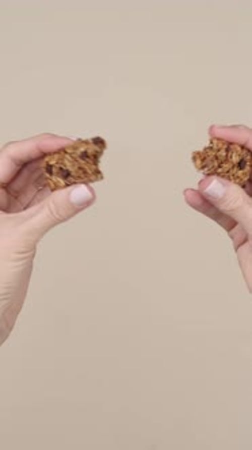 close-up of female hands with pink nails breaking an oat bar. The bar's crunchy texture and natural
