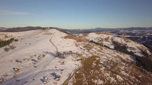 Luftaufnahme eines kleinen Dorfes mit verstreuten Häusern auf schneebedeckten Hügeln im Winter und kahler Dunkelheit