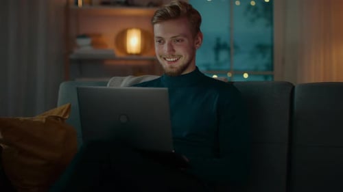 In the Evening Handsome Young Man at Home Sitting on a Couch in the Living Room Works on a Laptop C