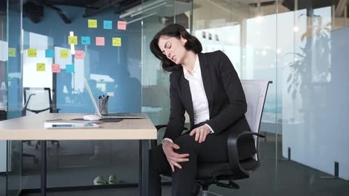 Businesswoman sitting in office feeling discomfort from knee pain. Workplace health and wellness