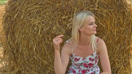 A happy adult woman in a summer dress rests in the shade of a straw bale in late summer