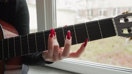 Close-up of a girl's hands playing chords on an acoustic guitar while sitting on a windowsill.