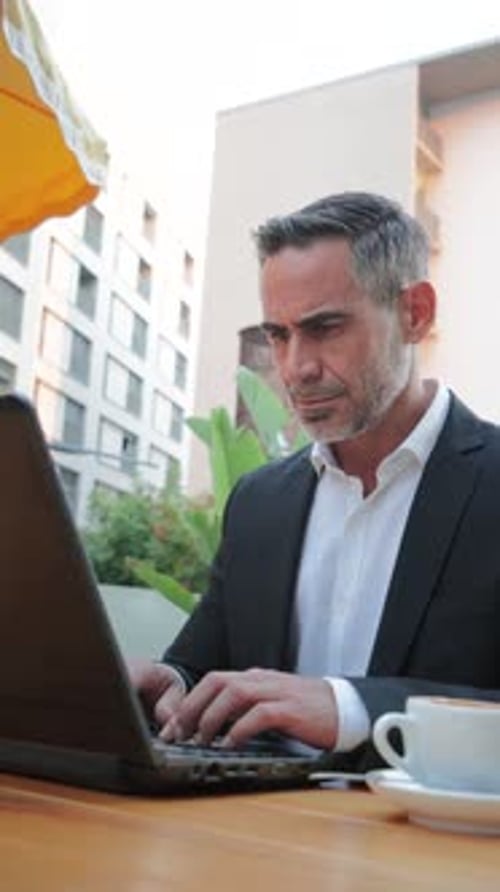 Focused Businessman in Suit Typing on Laptop While Enjoying Coffee at Outdoor Cafe During Afternoon