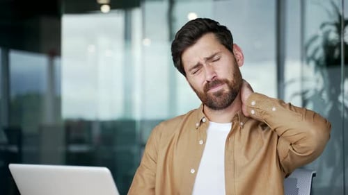Tired businessman suffering from neck pain while sitting at desk at workplace in business office.
