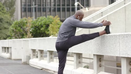 African American man lifting and stretching right leg on railing in plaza for warmup, copy space