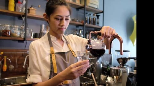 Young Asian barista woman in apron holds a drip coffee syphon and pours coffee in a mug.