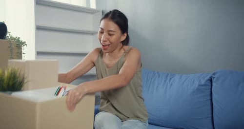 Asian woman in casual sitting on coach enjoy unpacking box stuff real estate purchase.