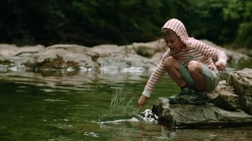 Little Boy Playing Near Cold Stream and Beating Water Surface with a Stick Creative Hiking in Wild