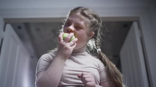 A Girl with Down Syndrome is Biting a Green Apple Healthy Food Disabled Person at Home Life with a