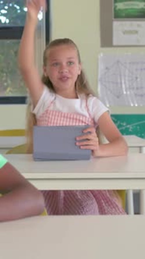 Vertical video: Raising hand, girl in pink dress answering teacher prompt in class, holding tablet