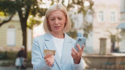 Frustrated Mature Businesswoman in Suit Using Credit Card and Mobile Phone Standing on City Street