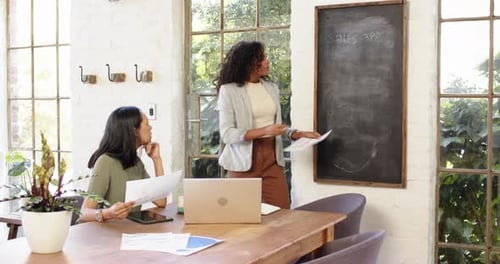 Multiracial female colleagues discussing sales strategy with laptop and charts in modern home office
