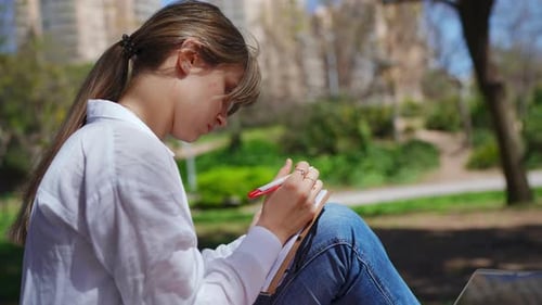 Focused Female Writer Working in Notebook with Red Pen Sitting on Park Bench Amid Urban Greenery
