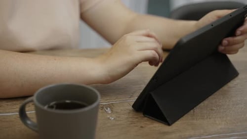Woman Taps on Screen of Tablet on Wooden Table with Cup Closeup