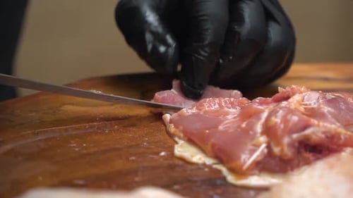 Um chef profissional cortando carne de frango crua em uma tábua de cortar de madeira usando luvas pretas.