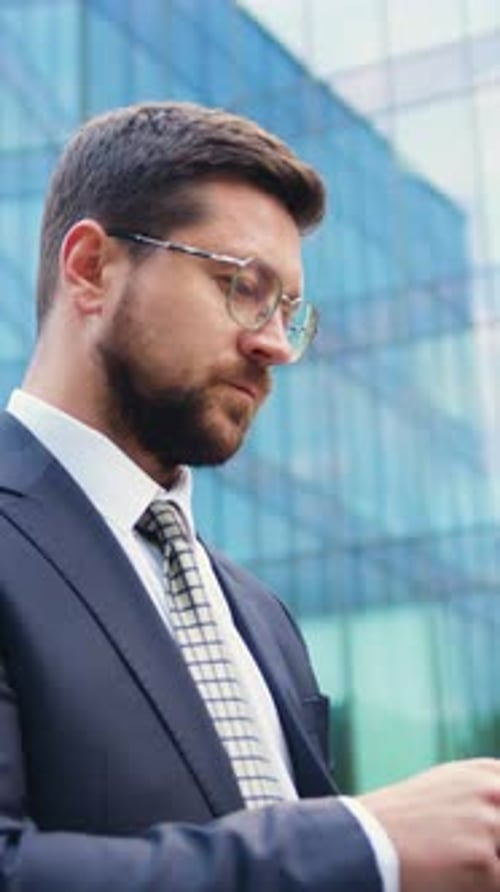 Side View of a Young Caucasian Man in Formal Clothes Walking on the Street While Using a Tablet with
