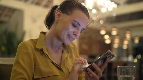 Young woman texting on smartphone and drinking water in cafe at night