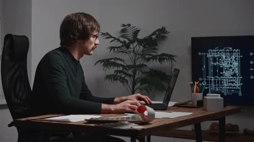An Architect Works on a Laptop with Blueprints in a Specialized Program