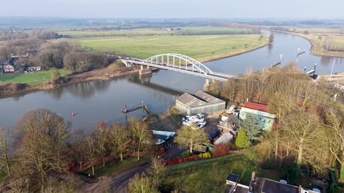 Aerial view of river with arched bridge linking wooded and industrial banks. Boats, buildings