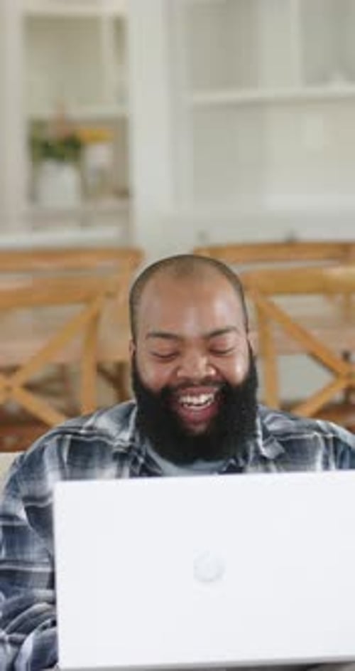 Vertical video: African American man in plaid reacting to white laptop clip laughing at home table