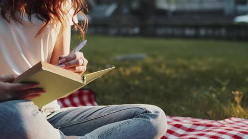 Young Woman in Casual Clothes Close Up Sitting on a Plaid in a City Park on a Summer and Writing in