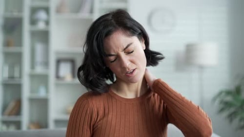 Tired young adult woman suffering from neck pain while sitting on sofa in the living room at home.