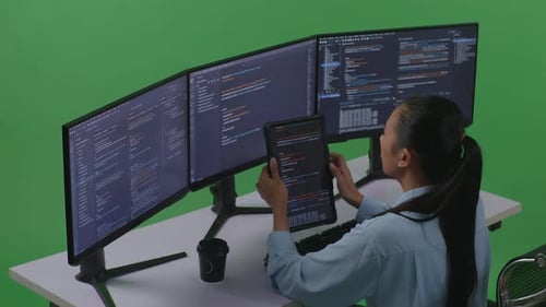 Side View Of Asian Woman Working With Tablet While Write Code With Multiple Computer In Green Screen
