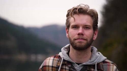 A young bearded man with wavy hair filmed in slow motion walks between hills and forest along a smal