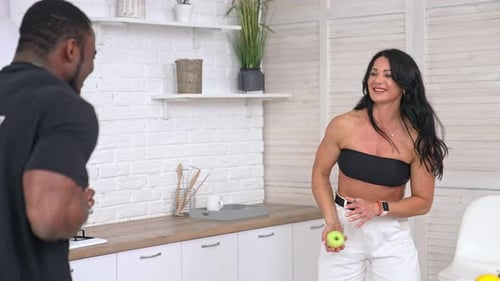 Cheerful mixed race couple throwing green apples to each other in the kitchen.