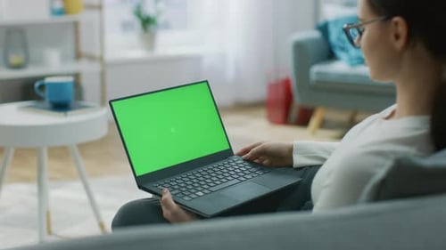 Young Woman at Home Sitting on a Couch Works on a Laptop Computer with Green Mock-up Screen. Girl U