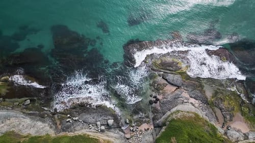 Aerial Topdown View of Turquoise Sea Waves Crashing Over Rocky Coastline with Moss and Clear Water
