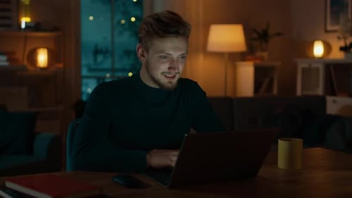 Handsome Smiling Man Works on a Laptop Computer while Sitting at His Desk at Home. Cozy Living Room