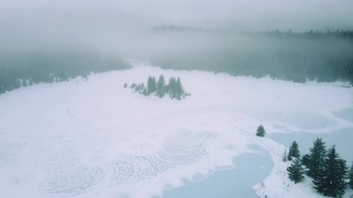 Aerial Of Snowy Frozen Lake With Moody Fog Clouds In Forest Trees