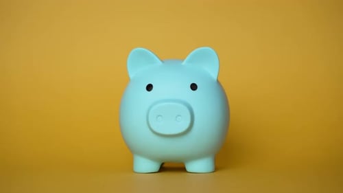 Hand Inserting Coins Into Light Blue Piggy Bank on Yellow Background