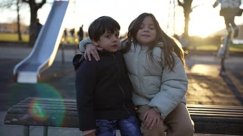 Little brother and sister seated at park bench during sunset time, backlight with flare. 5 yer old