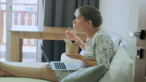 Side view of woman satisfying hunger with chinese udon noodles from paper wok sitting on the bed