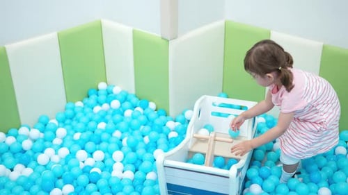 3-year-old Toddler Little Girl Plays in Dry Pool with White and Blue Plastic Balls, Throw Balls in W
