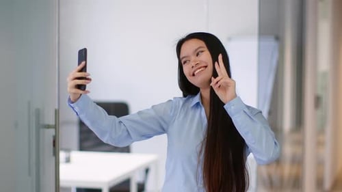 Young Asian Woman Taking a Selfie in an Office Space While Smiling and Posing With Her Finger