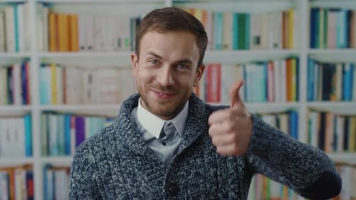 A Man in a Sweater and Tie Gives a Thumbs Up in a Cozy Library