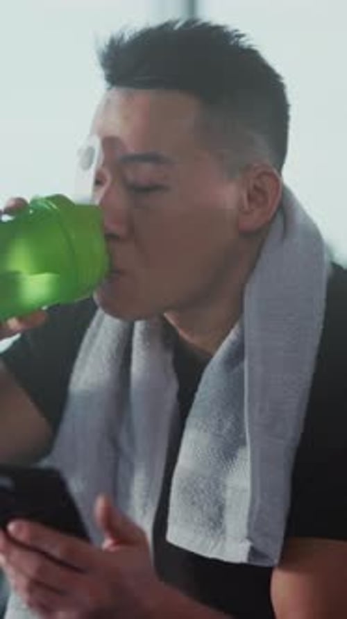 Athletic man drinking protein and using phone indoors